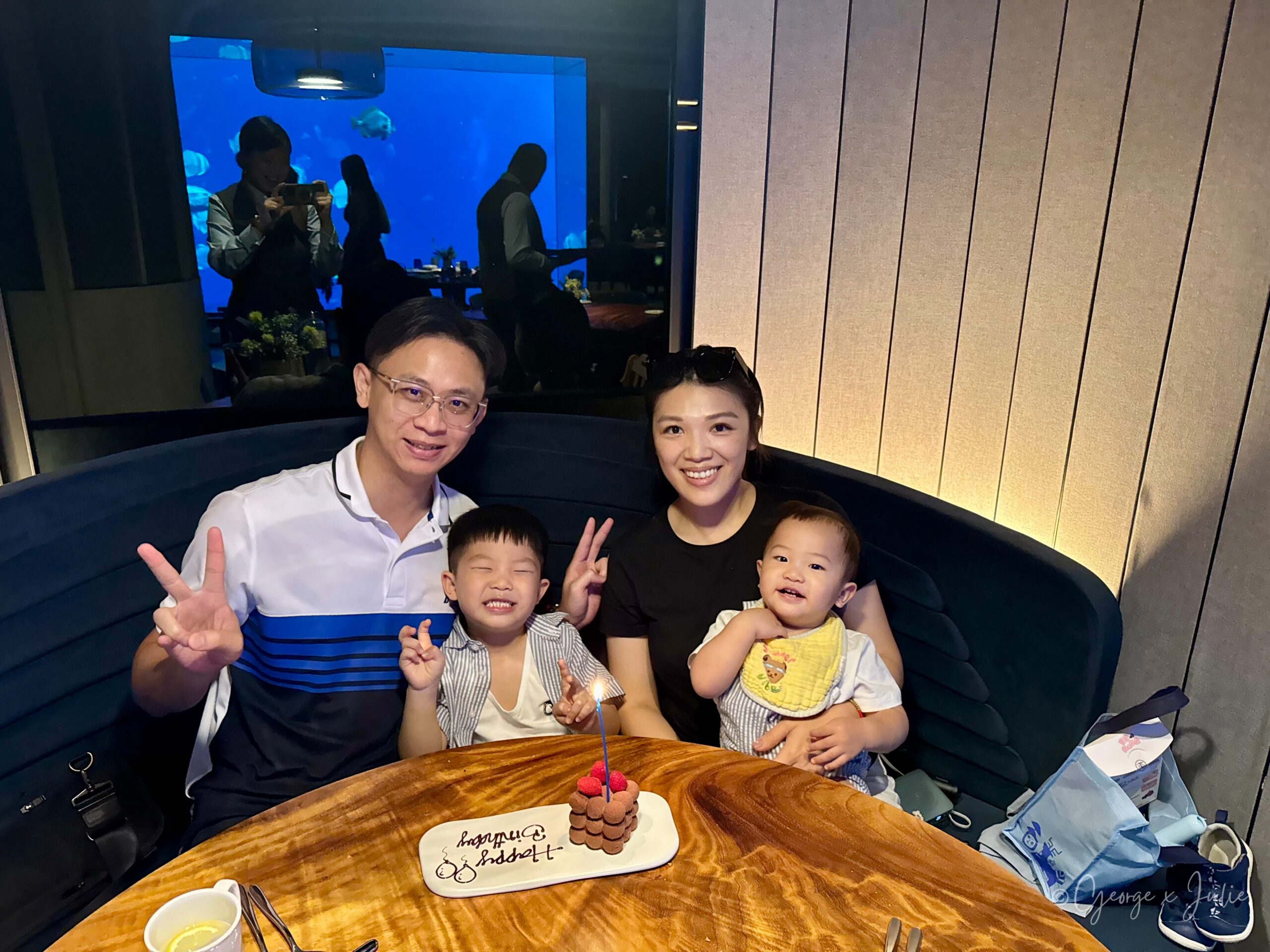 George、Julie和兩個孩子在桌前合照，桌上擺著插了蠟燭的巧克力生日蛋糕，背景是水族箱