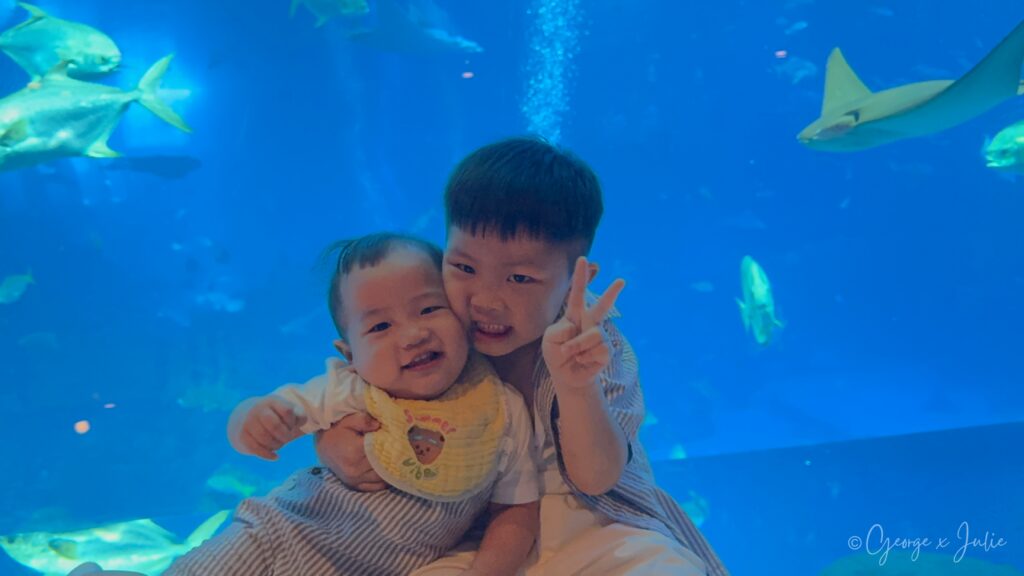 兩個孩子在水族箱前擁抱合照，背景魚群悠游