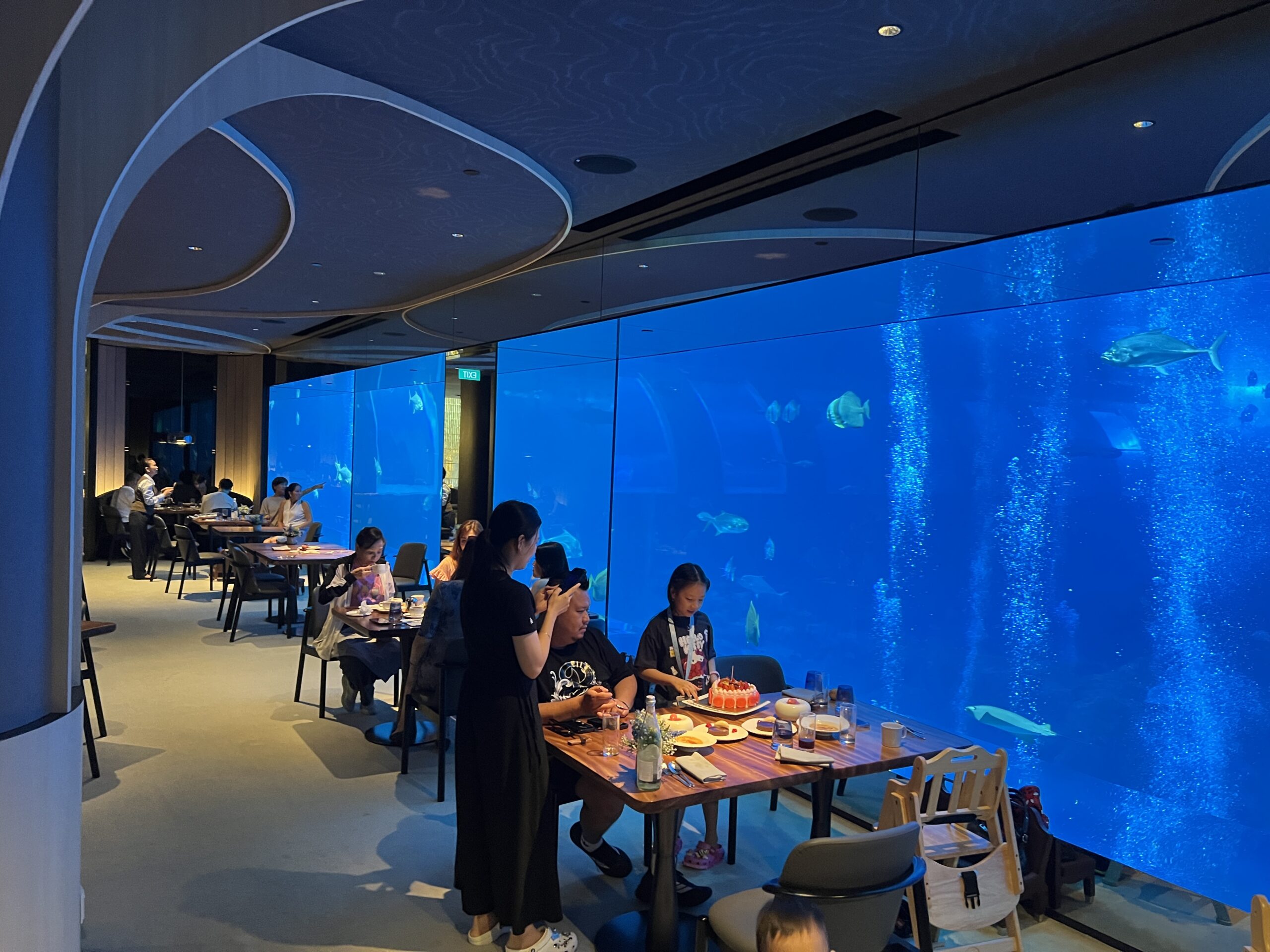餐廳環景，桌上擺著生日蛋糕，店員在一旁幫忙拍照留念，背景是整排水族箱與其他用餐客人