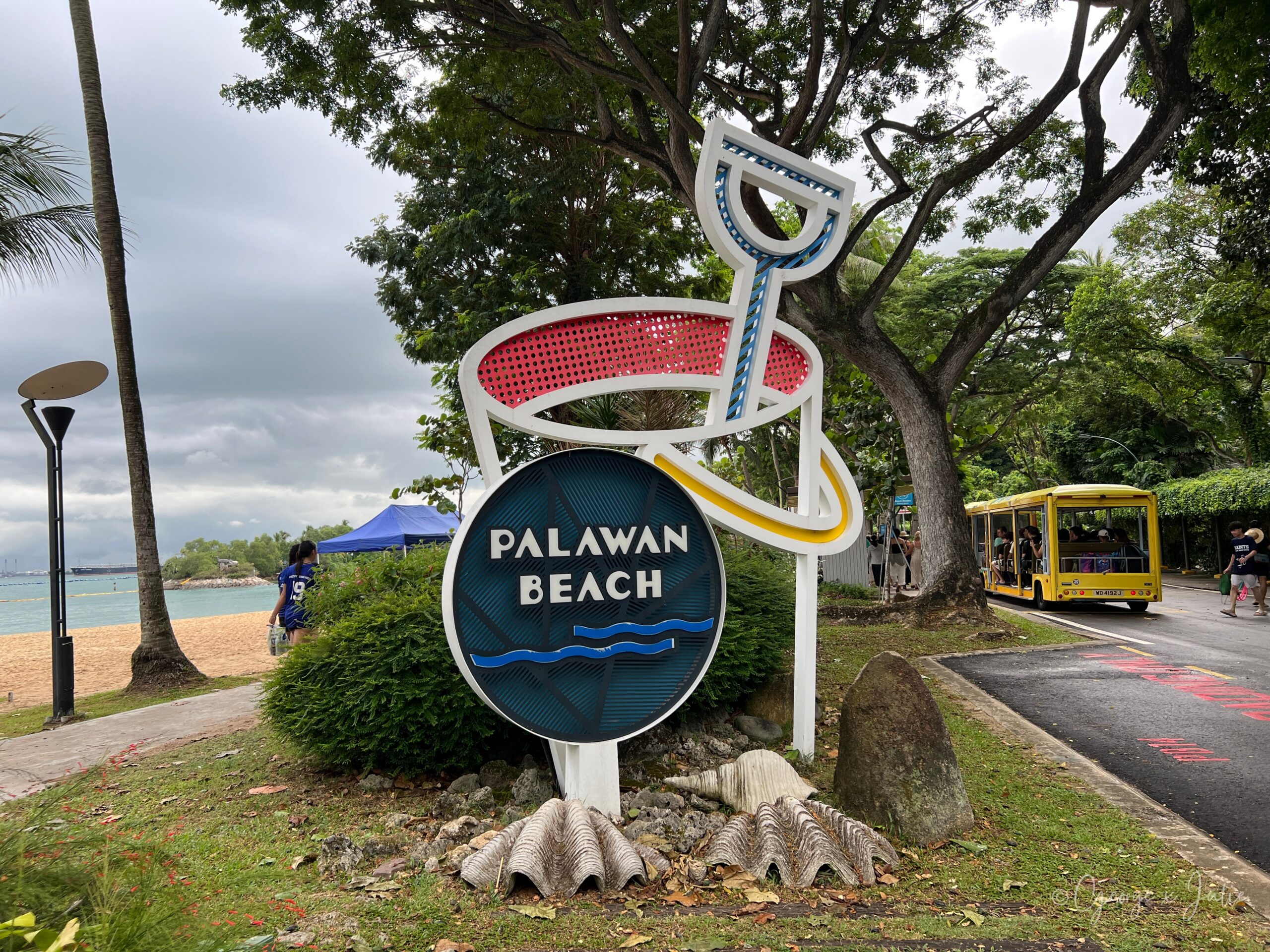 【新加坡景點】聖淘沙亞洲大陸最南端|Palawan Beach 90度椰子樹、吊橋免費踩點 2 PALAWAN BEACH 招牌特寫,貝殼與海浪圖案裝飾,後方一輛黃色接駁車正經過