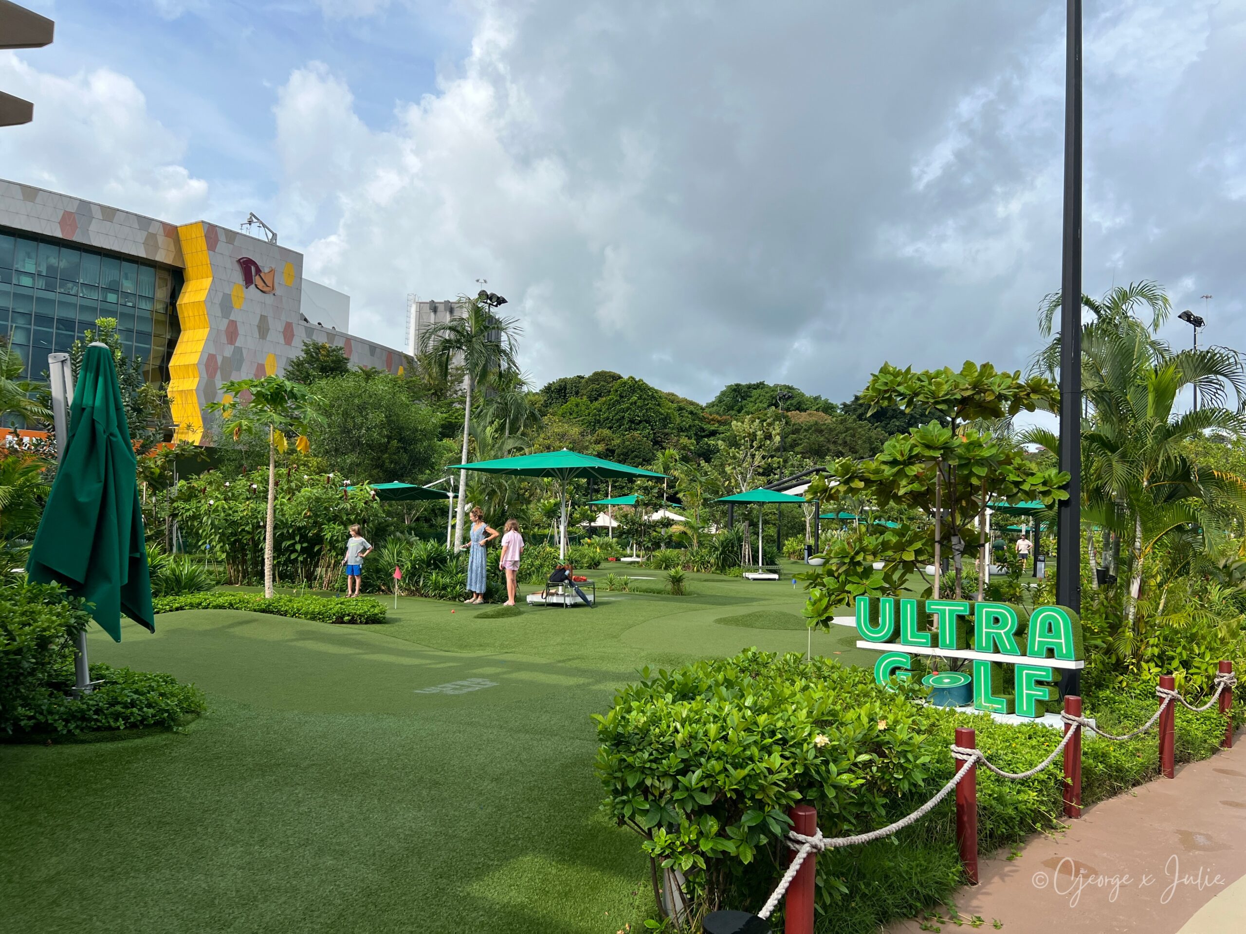 從入口旁望向整個 UltraGolf 球場，綠色大陽傘和高低起伏的果嶺，遠處有其他遊客在打球，右邊是 ULTRA GOLF 招牌