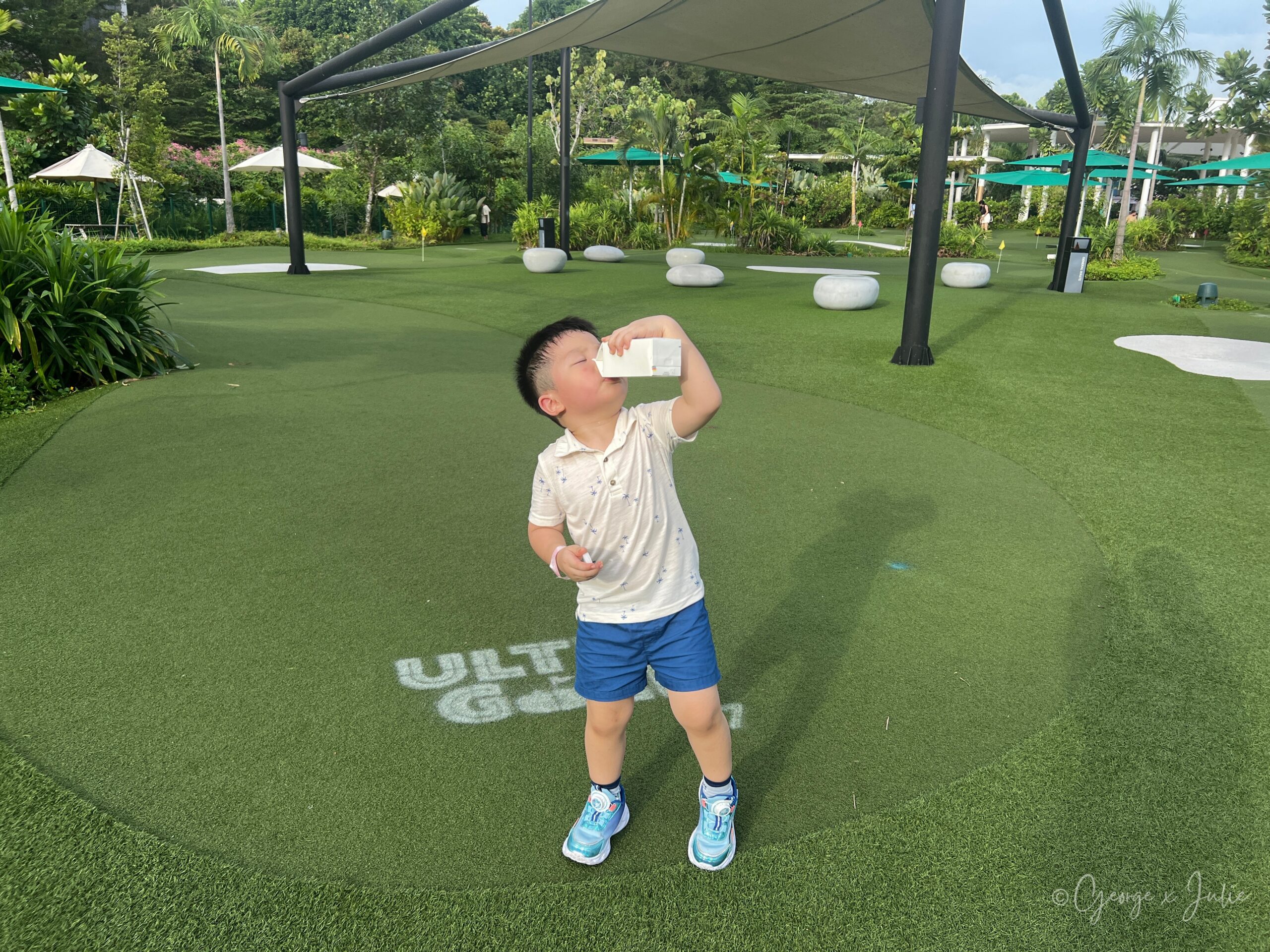 Jensen在球道邊的遮陽棚下站著喝飲料，腳邊的草皮上印著ULTRA GOLF字樣，後方有幾張白色圓石造景座位