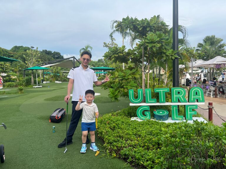 UltraGolf聖淘沙：George和Jensen各拿著推桿，在 ULTRA GOLF 招牌旁一起比出勝利手勢，背景是一整片球道
