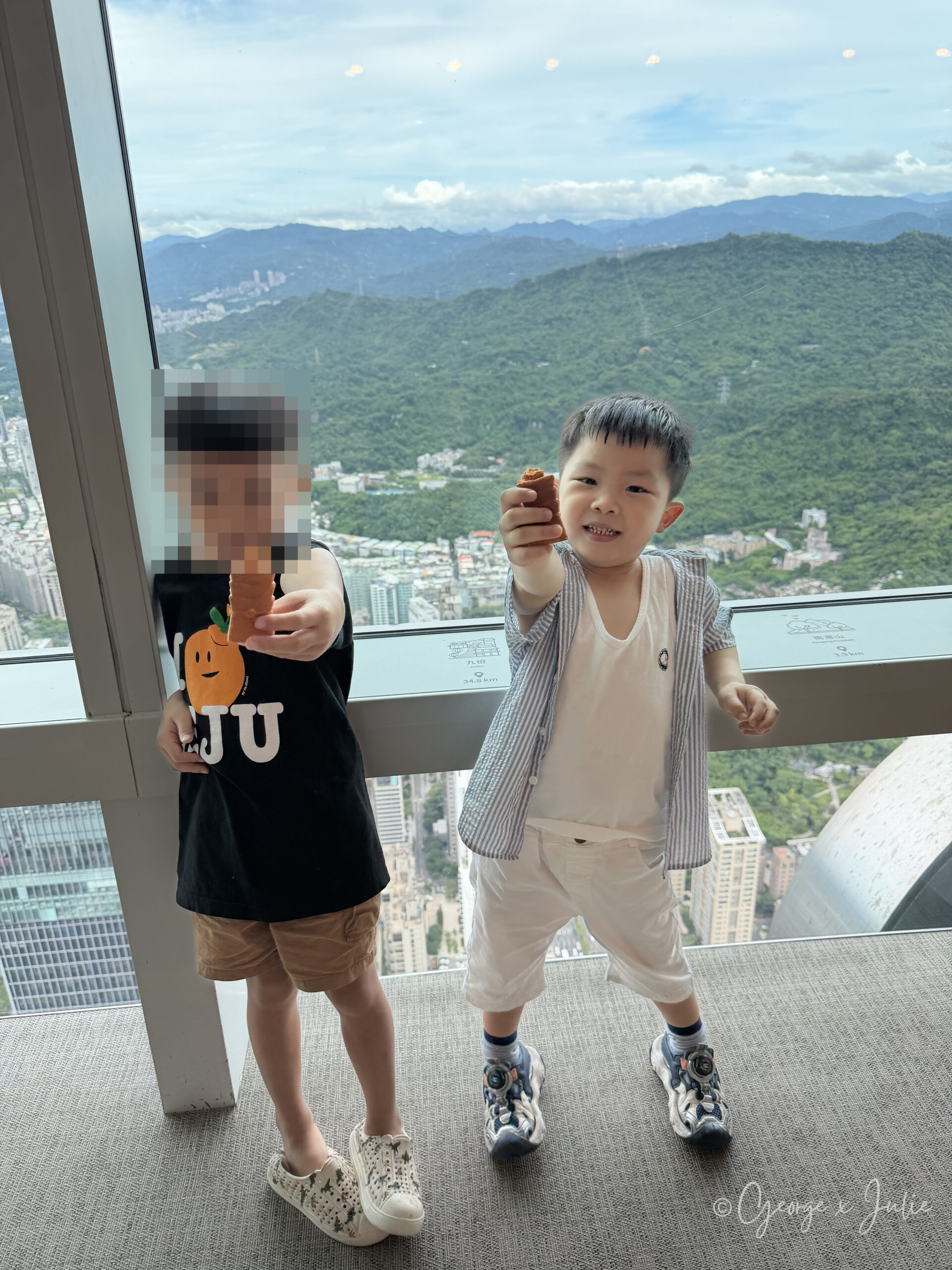 兩個小朋友站在台北101觀景台窗邊，拿著雞蛋糕對著鏡頭