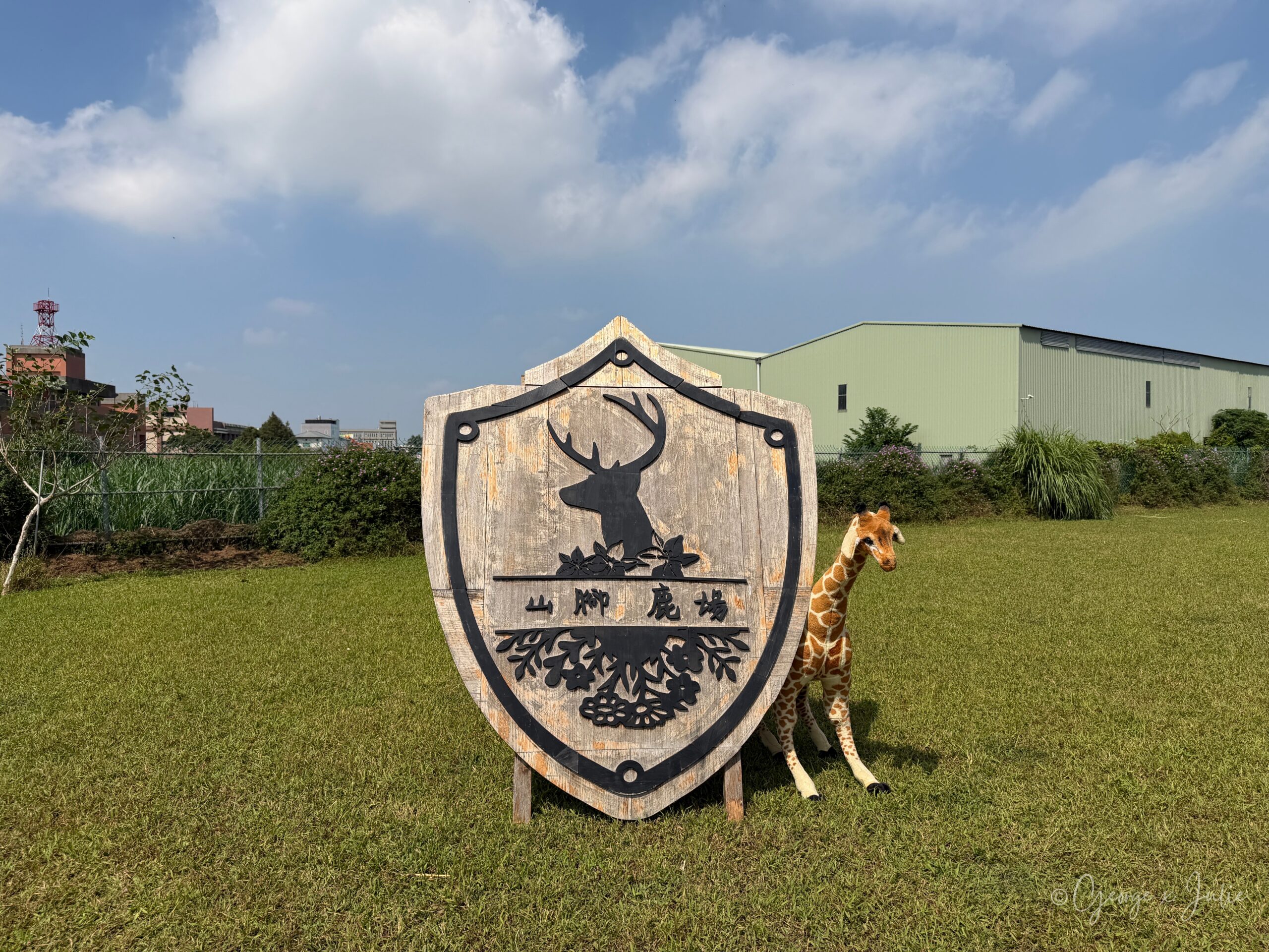 山腳鹿場木頭盾牌招牌，旁邊站著長頸鹿玩偶，背景是草地